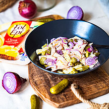 Schweizer Wurstsalat in einer schwarzen Schüssel auf einem rustikalem Holzbrett. Garniert mit Gewürzgurken und angeschnittenen Zwiebeln.