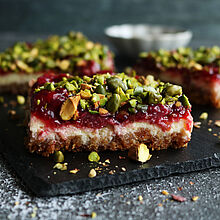 Kirsch-Pistazien-Cheesecake mit knusprigem Boden, fruchtigem Kirschbelag und gehackten Pistazien obendrauf, serviert auf einer dunklen Schieferplatte.