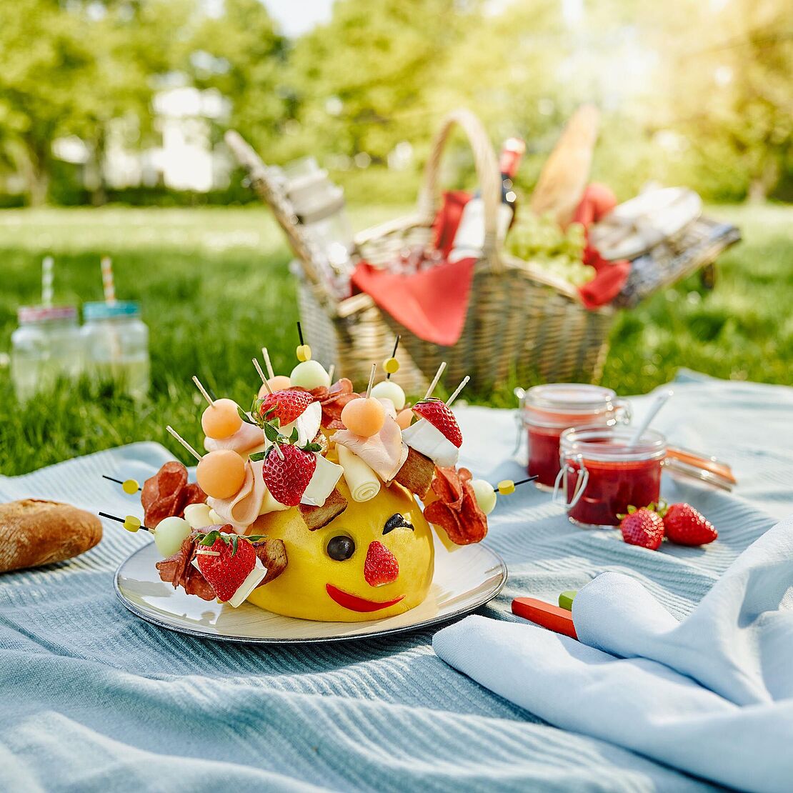 Käseigel Gérard hat auf seinen Spießen Géramont, Honigmelone und Putenbrust und sitzt auf einer Picknickdecke