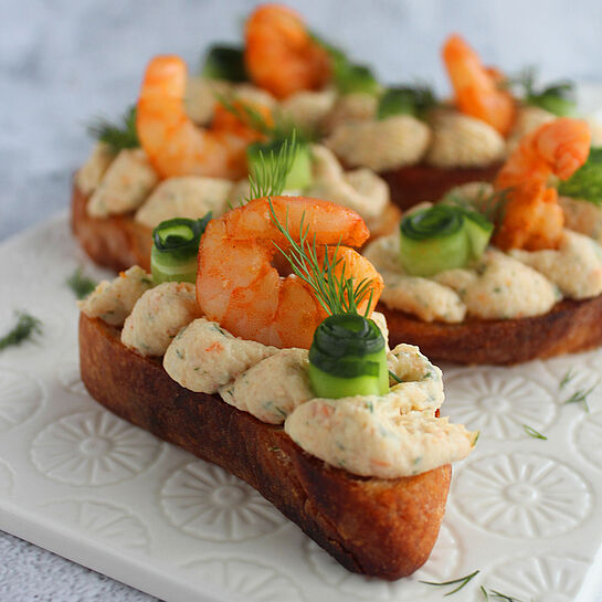 Canapés Rezept mit Schrimps, Gurke & Dill Elegante Canapés aus knusprigem Baguette mit marinierten Schrimps, cremiger Dill-Mousse und frischen Gurkenstreifen, dekorativ angerichtet.