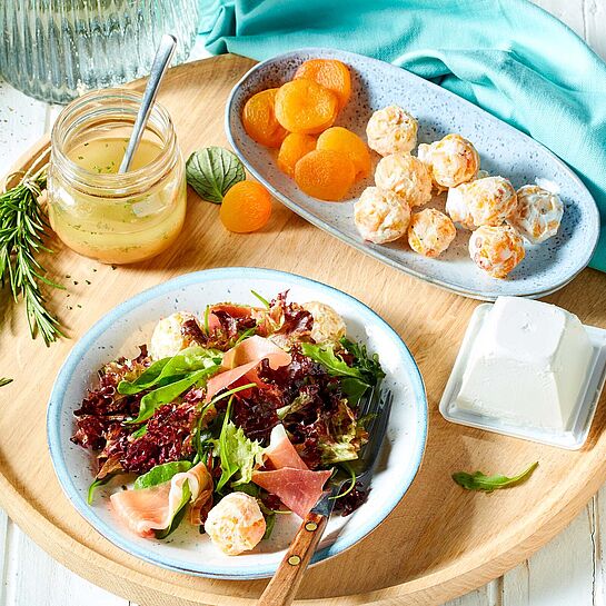 Holztablett mit einem leichten Sommersalat, der mit Aprikosen-Frischkäse-Bällchen und Serrano-Schinken garniert ist.