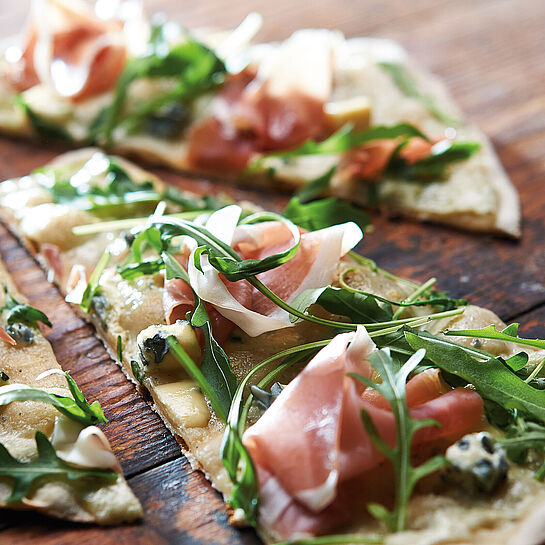 Dünner Flammkuchen mit Rucola und Bauernschinken auf einer rustikalen Holzunterlage 