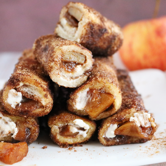 Apfel-Zimt-Toaströllchen mit Frischkäse Gestapelte Apfel-Zimt-Toaströllchen mit Frischkäsefüllung auf einem weißen Teller, gefüllt mit karamellisierten Apfelstücken und umhüllt von einer Zimt-Zucker-Kruste.