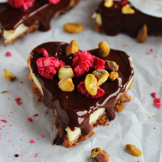 Cheesecake-Herzen mit Schokoladen-Topping und edler Garnitur Herzförmige Cheesecakes mit knusprigem Keksboden, cremiger Füllung und glänzendem Schokoladenüberzug, dekoriert mit Pistazien, Himbeeren und Kokoschips.